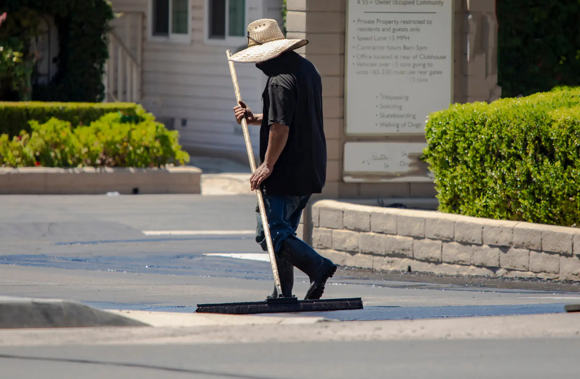 A person wearing a wide-brimmed hat and dark clothing uses a large squeegee to spread material on a paved surface, possibly sealing or cleaning it, near a landscaped area and buildings.