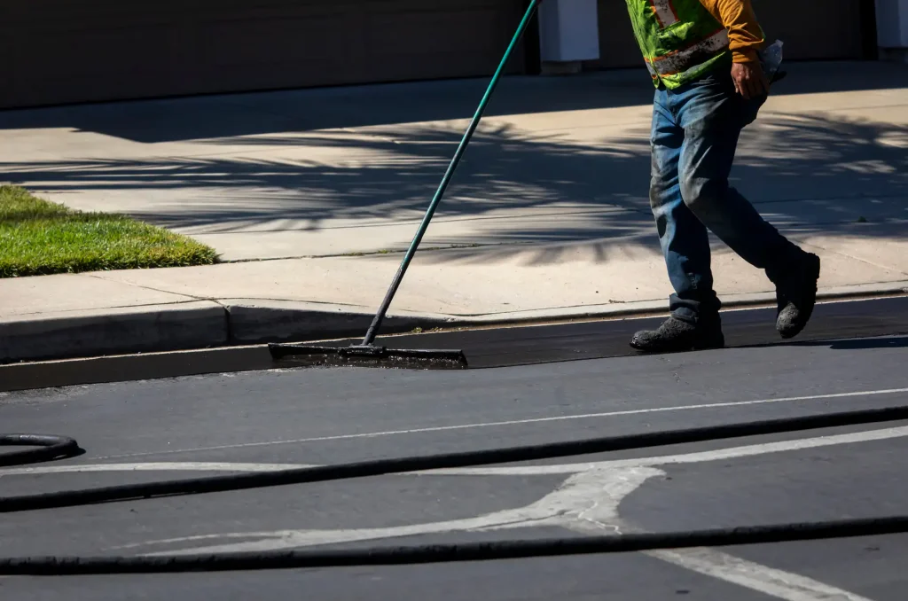 A worker wearing jeans and a safety vest uses a long-handled tool to smooth freshly laid asphalt on a street near a curb, with hoses laying across the road.