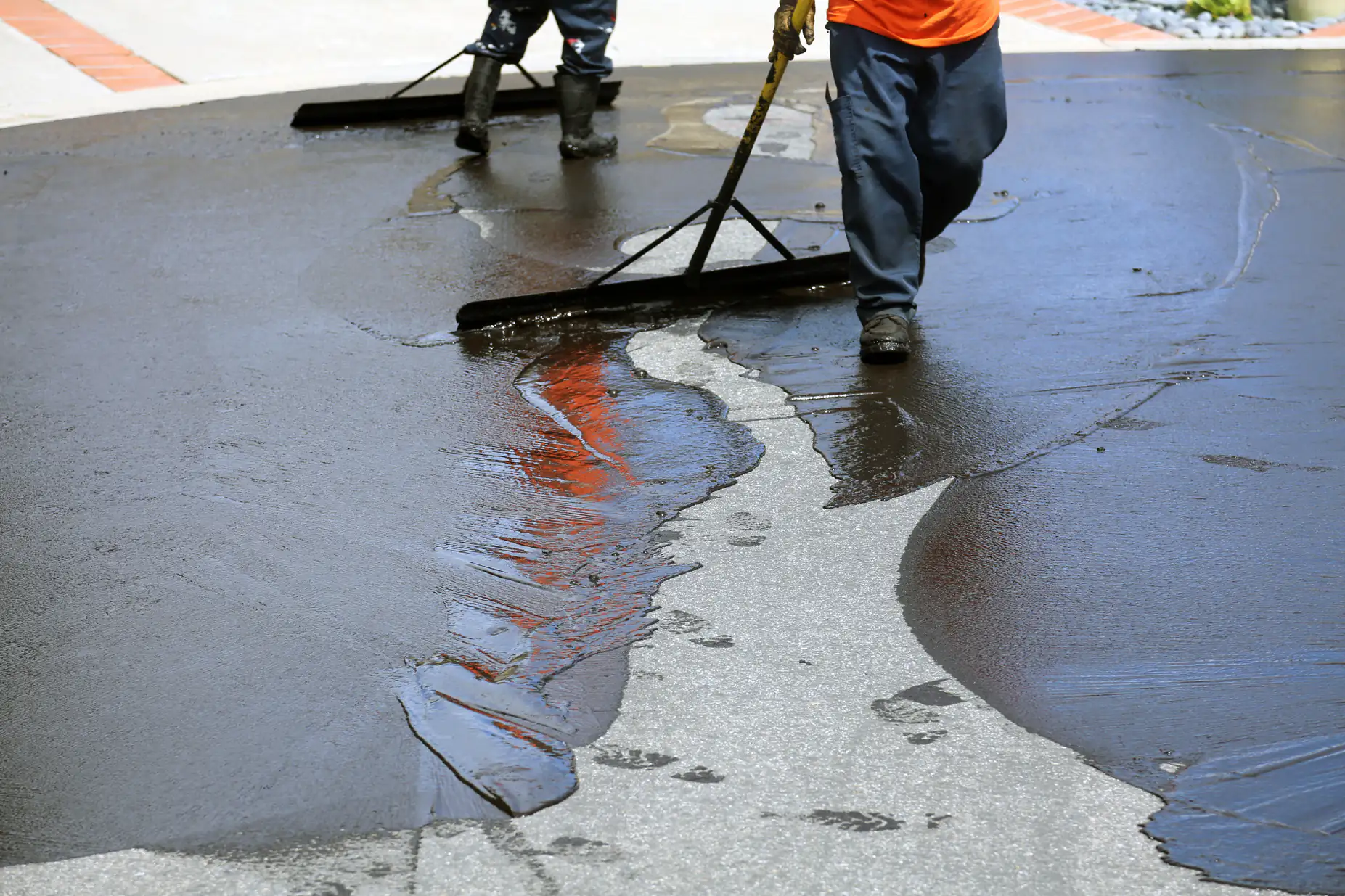 Two workers spread a fresh layer of asphalt or sealant on a road using large squeegees. Wet black surface contrasts with dry pavement, and footprints are visible in the newly applied material.