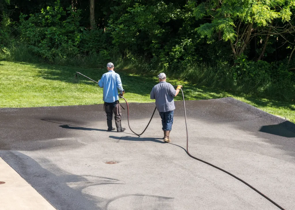 Two workers wearing hats apply liquid sealant with hoses to a blacktop driveway on a sunny day, with trees and green grass in the background.