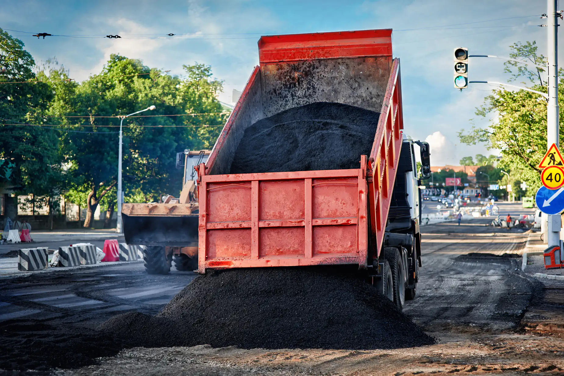 A red dump truck unloads black asphalt onto a city street under construction. A loader works nearby, with traffic signs, barriers, and trees visible in the background.