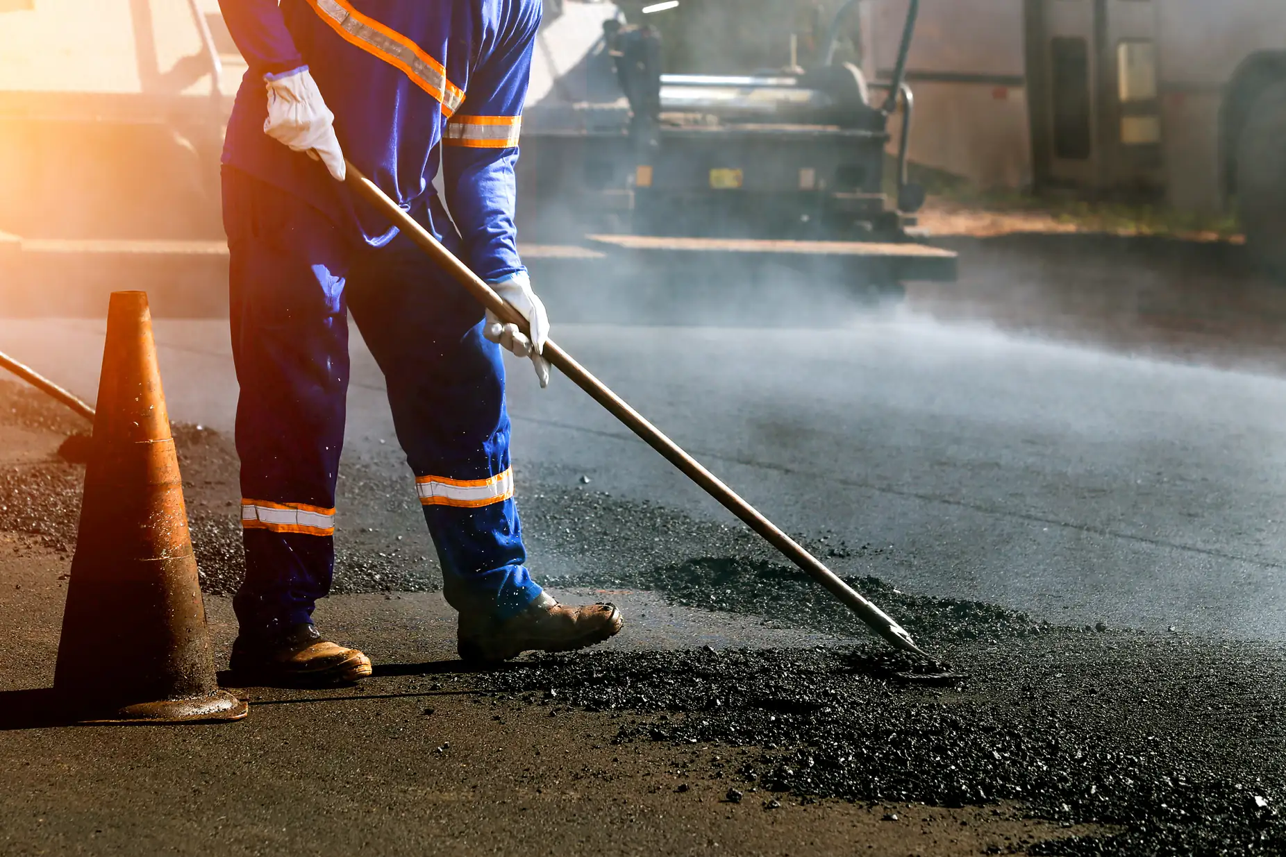 A worker in blue reflective clothing spreads asphalt on a road with a tool, near an orange traffic cone, while steam rises from the freshly laid surface.