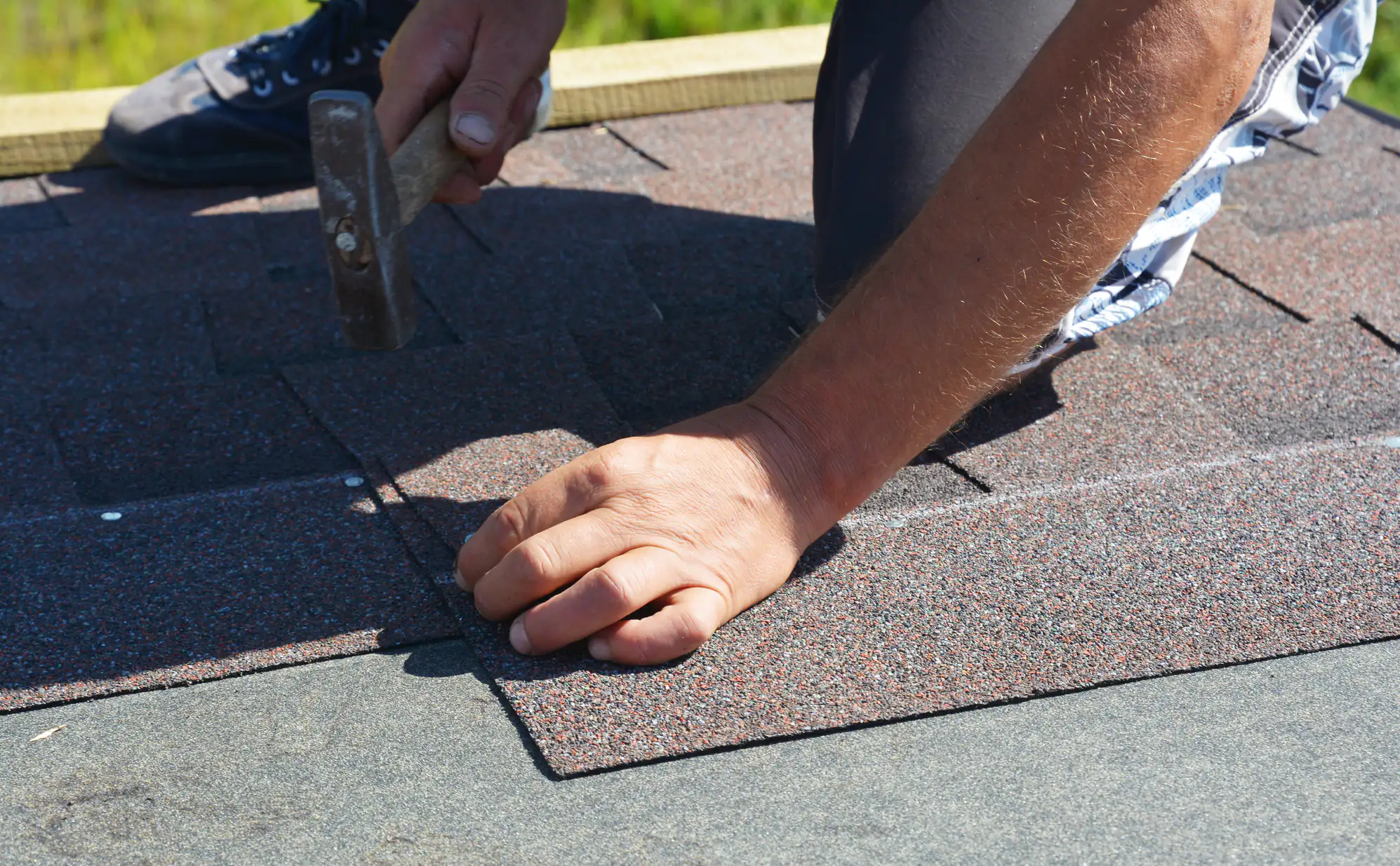 A person is installing asphalt roof shingles, using a hammer to secure the shingles in place on a rooftop under bright sunlight. Only the person's hands and part of their legs are visible.