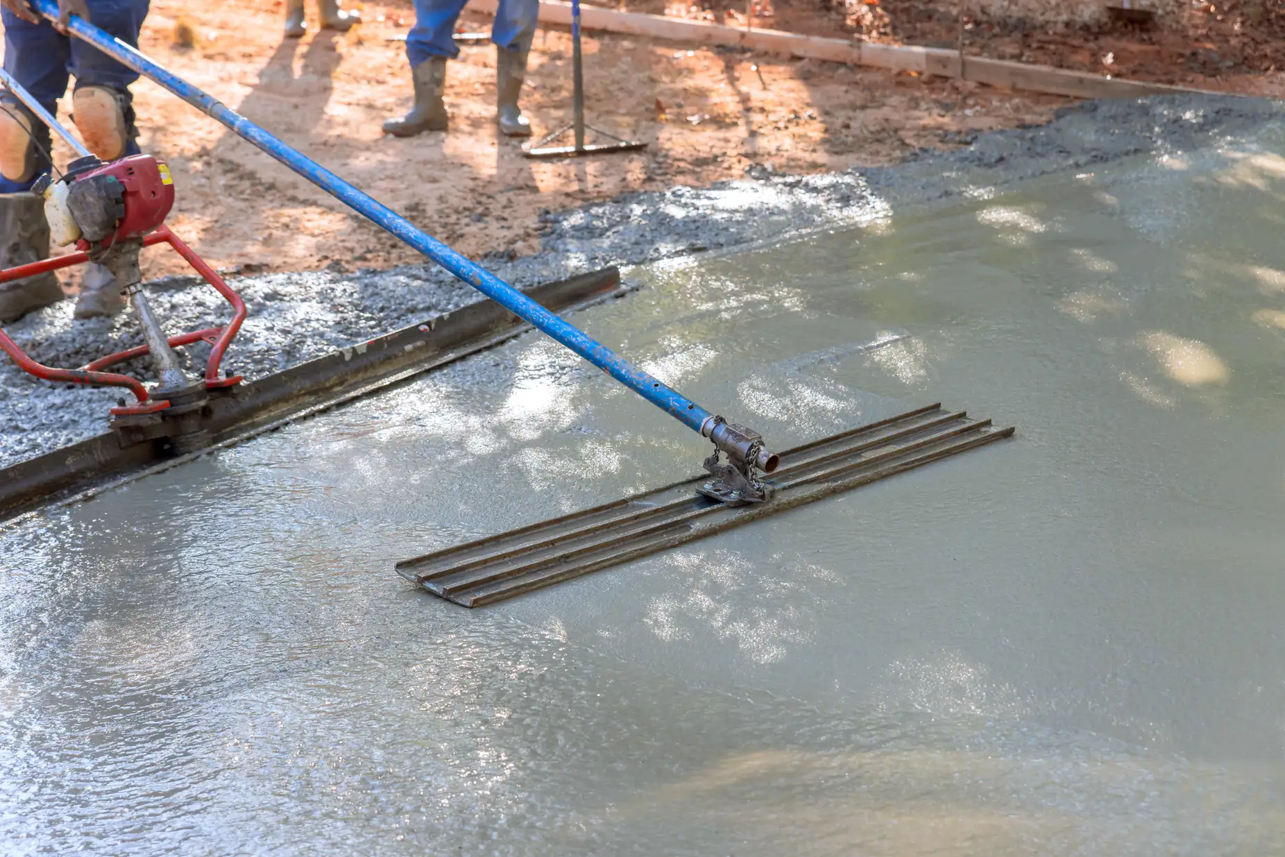 A close-up of workers using a large blue bull float to smooth and level freshly poured concrete at a construction site. The wet concrete reflects light, and workers' legs are visible in the background.