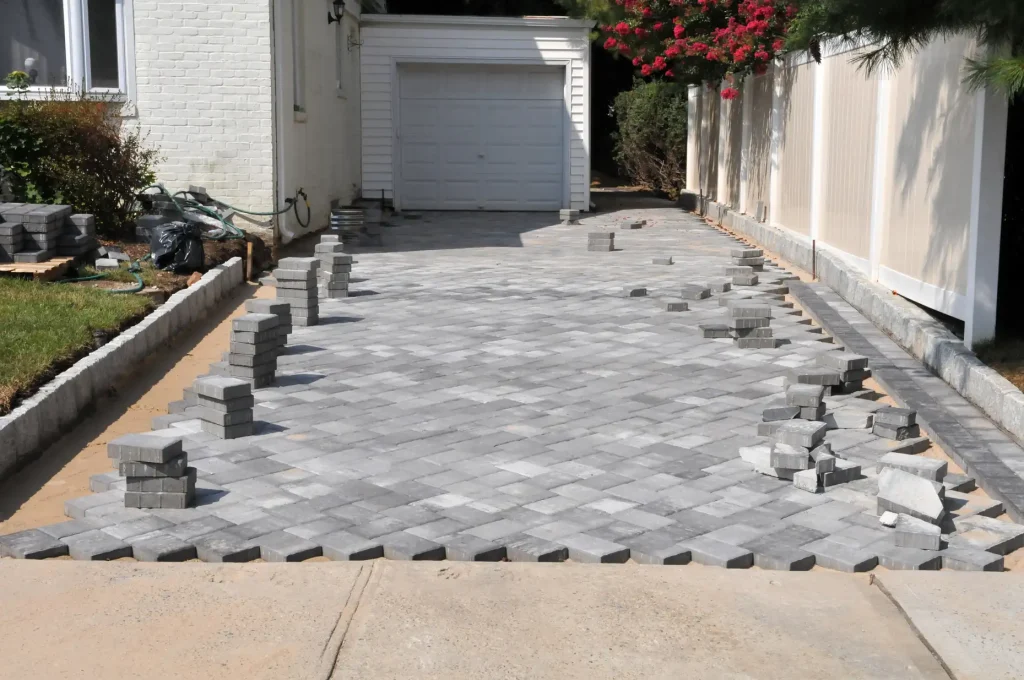 A driveway is under construction with gray pavers arranged in a herringbone pattern. Stacks of unused pavers are placed along the edges, and a garage is visible at the end of the driveway.