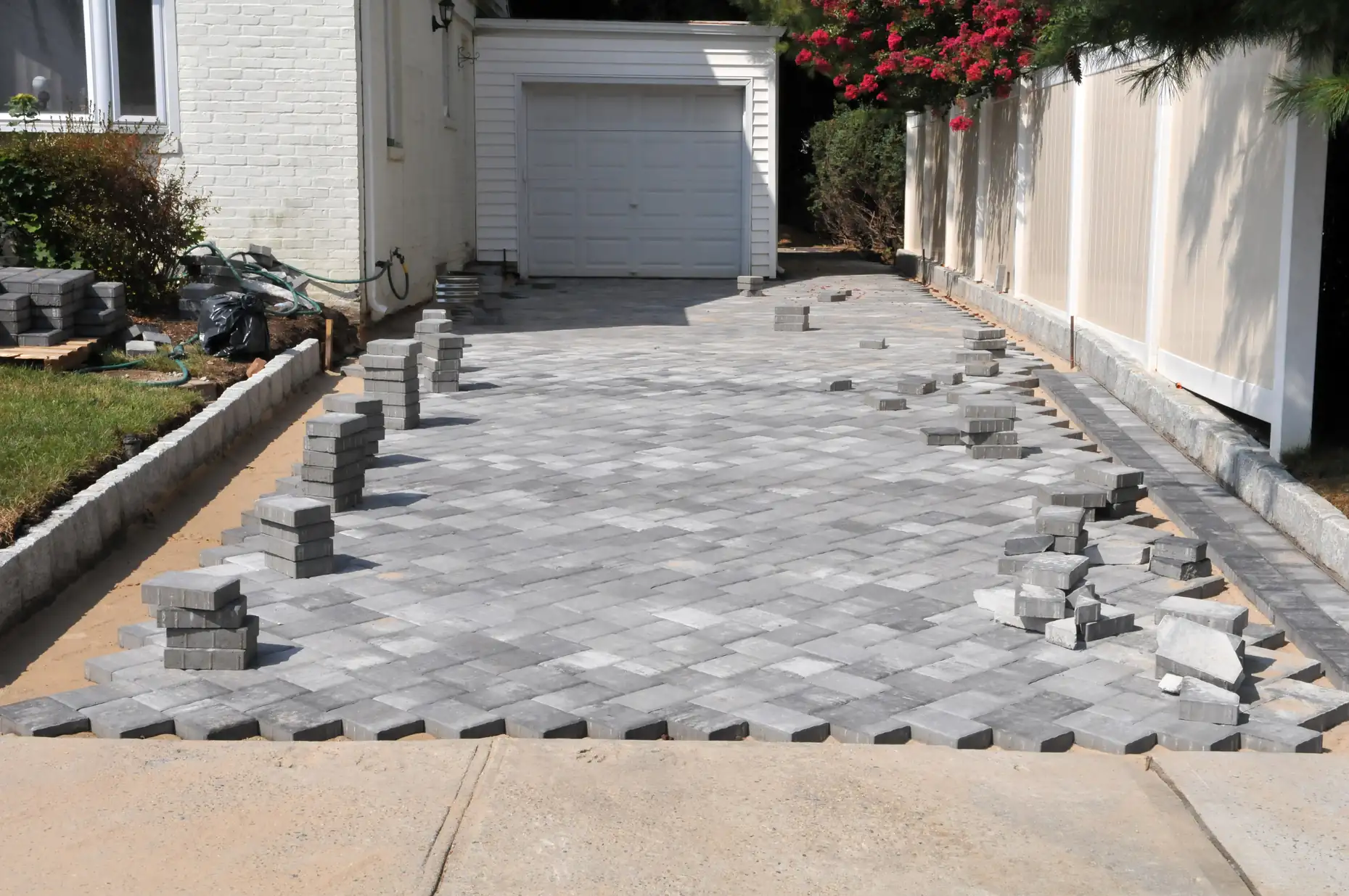 A driveway is under construction with gray pavers arranged in a herringbone pattern. Stacks of unused pavers are placed along the edges, and a garage is visible at the end of the driveway.