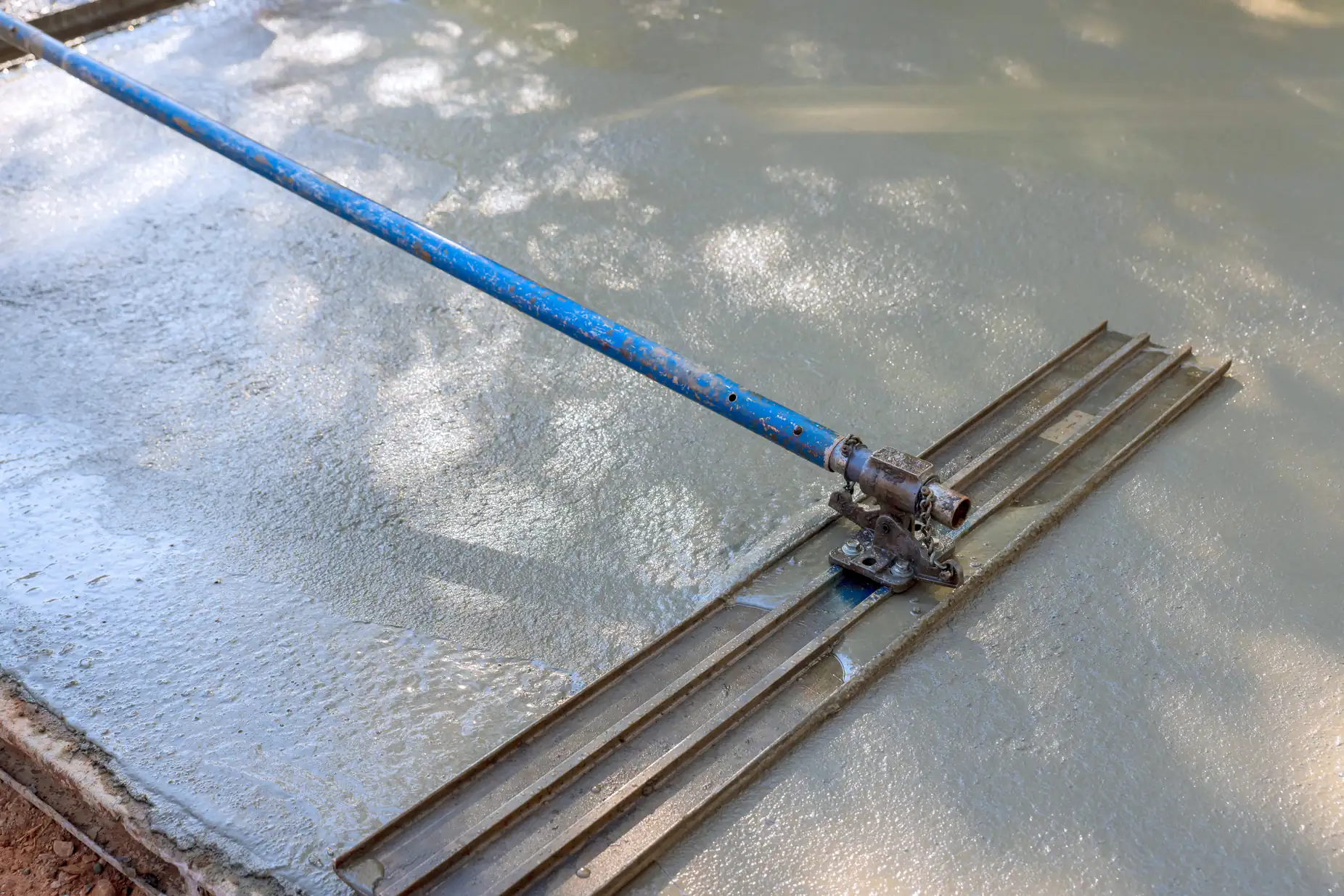 A blue bull float is being used to smooth and level freshly poured concrete, creating an even surface. Sunlight and shadows are visible on the wet concrete.