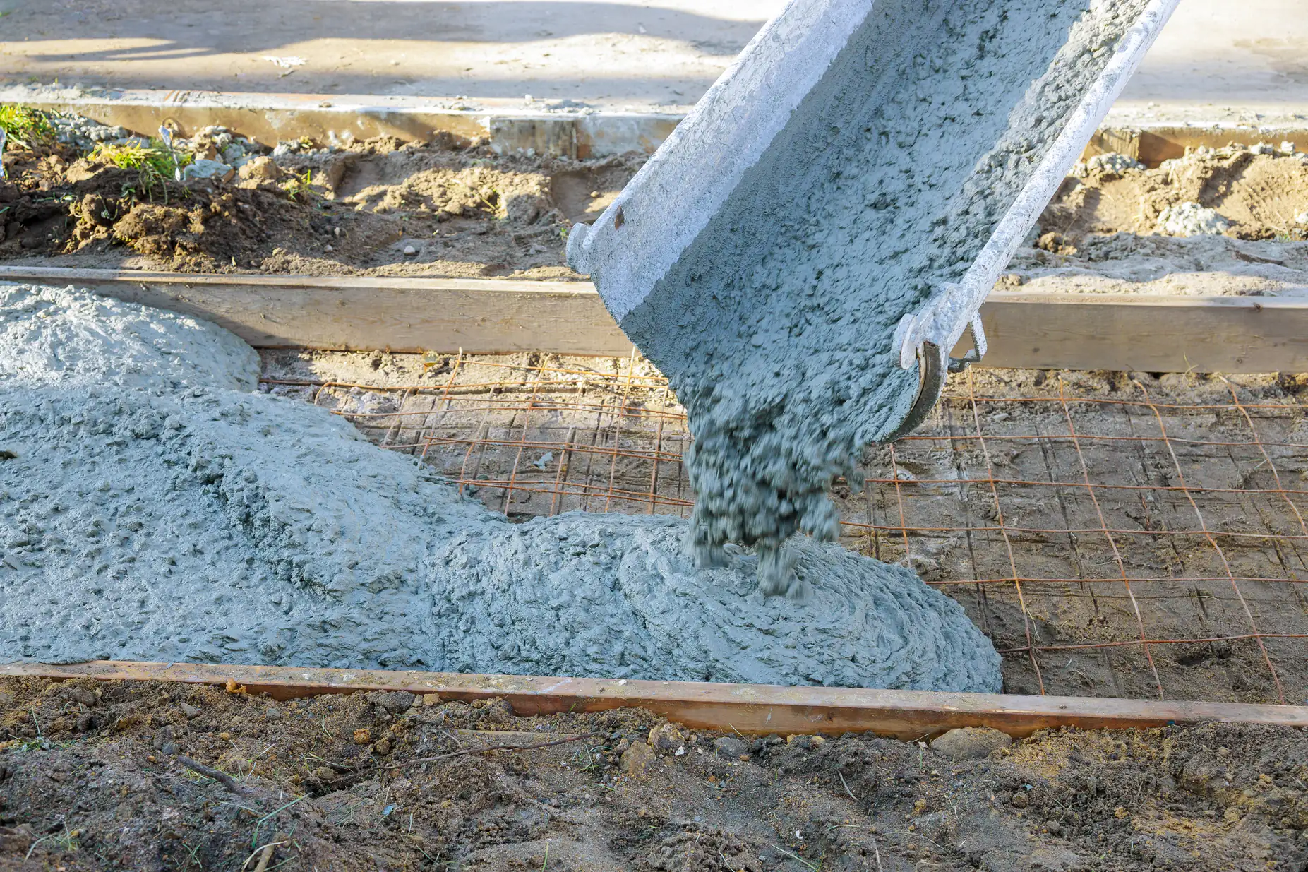 Wet concrete is being poured from a chute onto a prepared area with wire mesh and wooden framing, forming the base for a new pavement or slab. The surroundings include soil and construction materials.