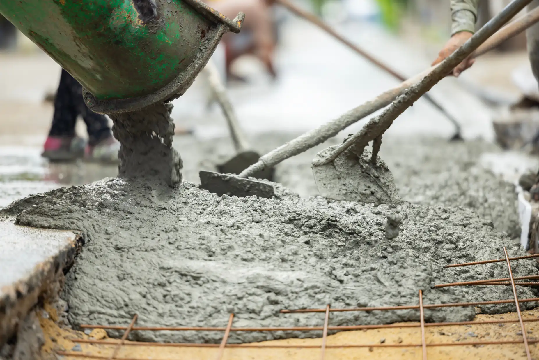 Workers pour and spread wet concrete from a mixer onto a construction site, using shovels to level the surface over exposed rebar.