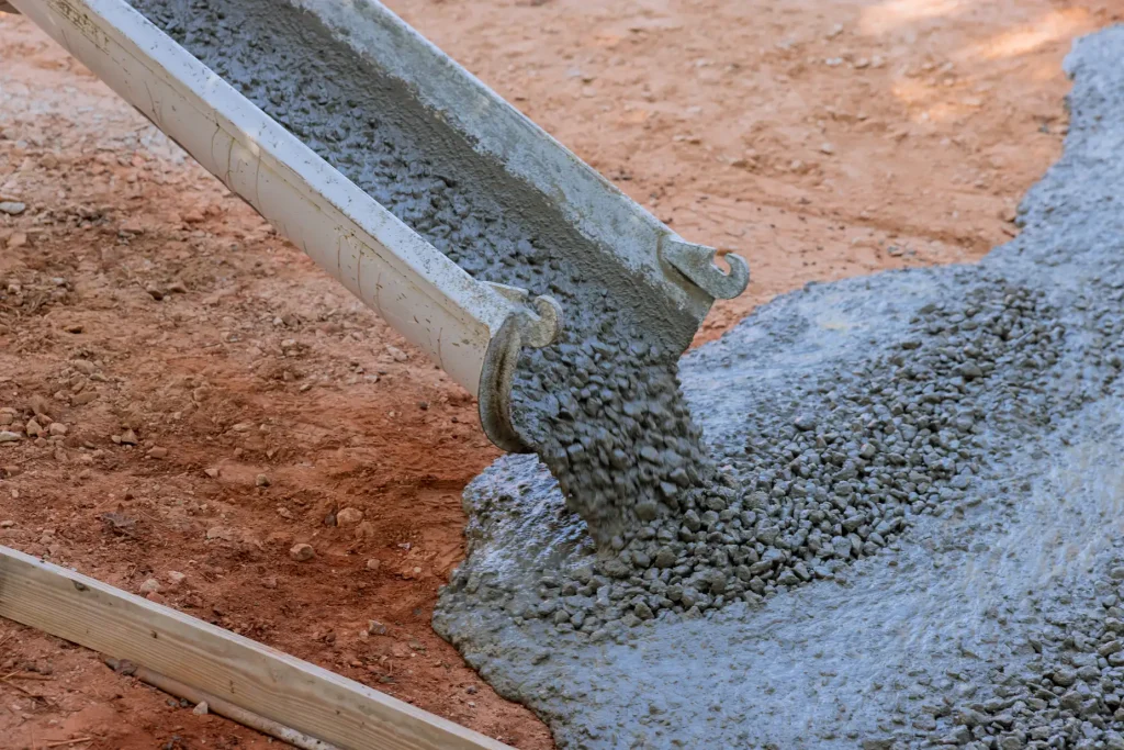 Wet concrete mixture is being poured from a metal chute onto a reddish dirt ground at a construction site, with a wooden board visible in the corner for formwork.