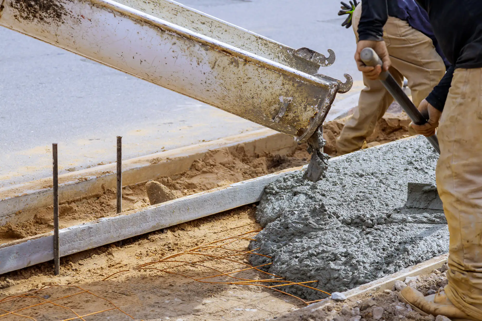 Wet concrete is being poured from a chute onto a prepared area with metal rebar, as construction workers guide and smooth the mixture to form a sidewalk or curb.