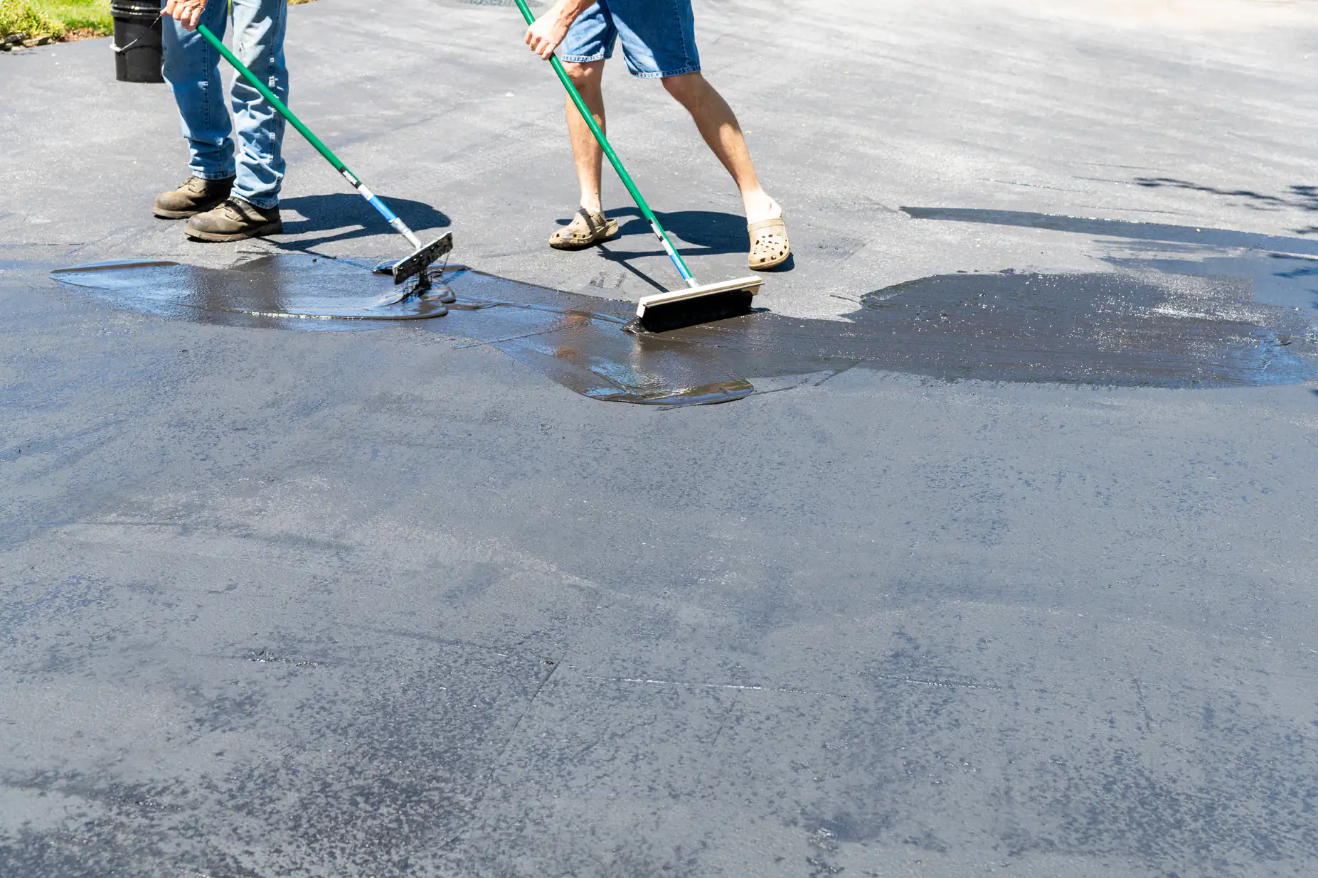 Two people wearing shorts and jeans use long-handled brushes to spread black sealant on a driveway under bright sunlight. The surface appears shiny and wet where the sealant has been applied.
