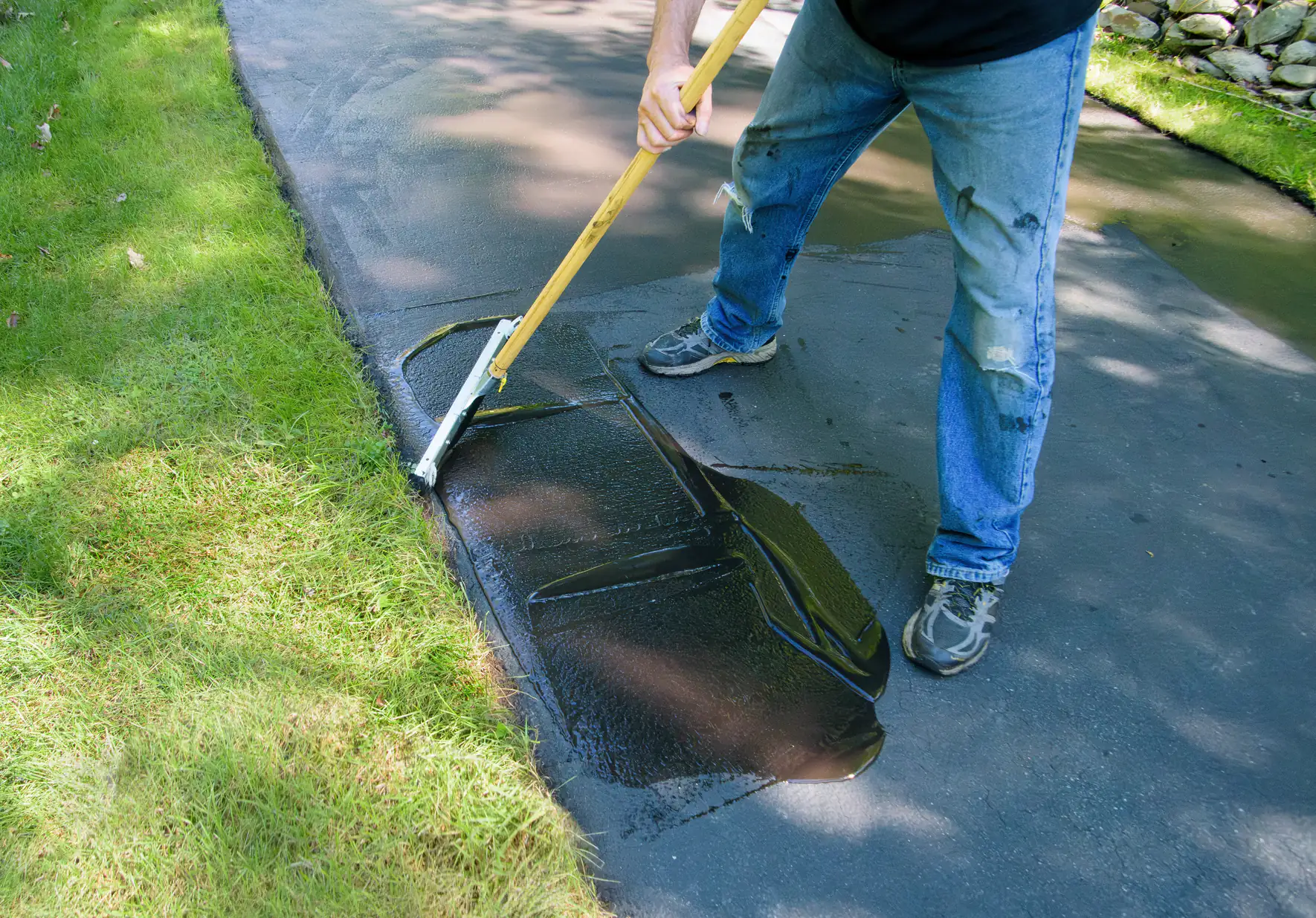 A person in ripped jeans uses a long-handled tool to spread black sealant on a driveway, with green grass along the edge and rocks visible in the background.