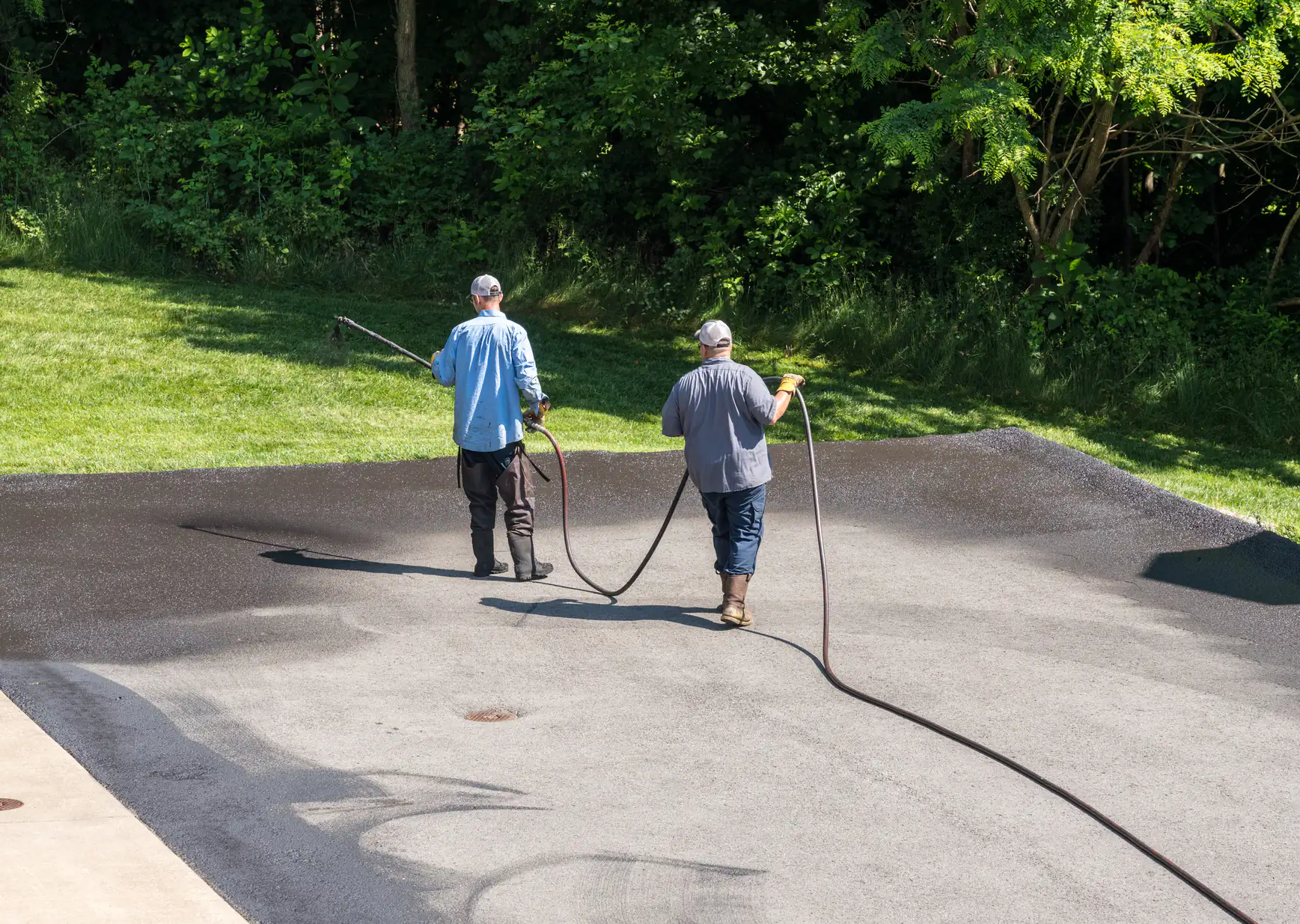 Two workers in casual clothing and hats are applying a dark sealant to a driveway with hoses, with green grass and trees in the background.