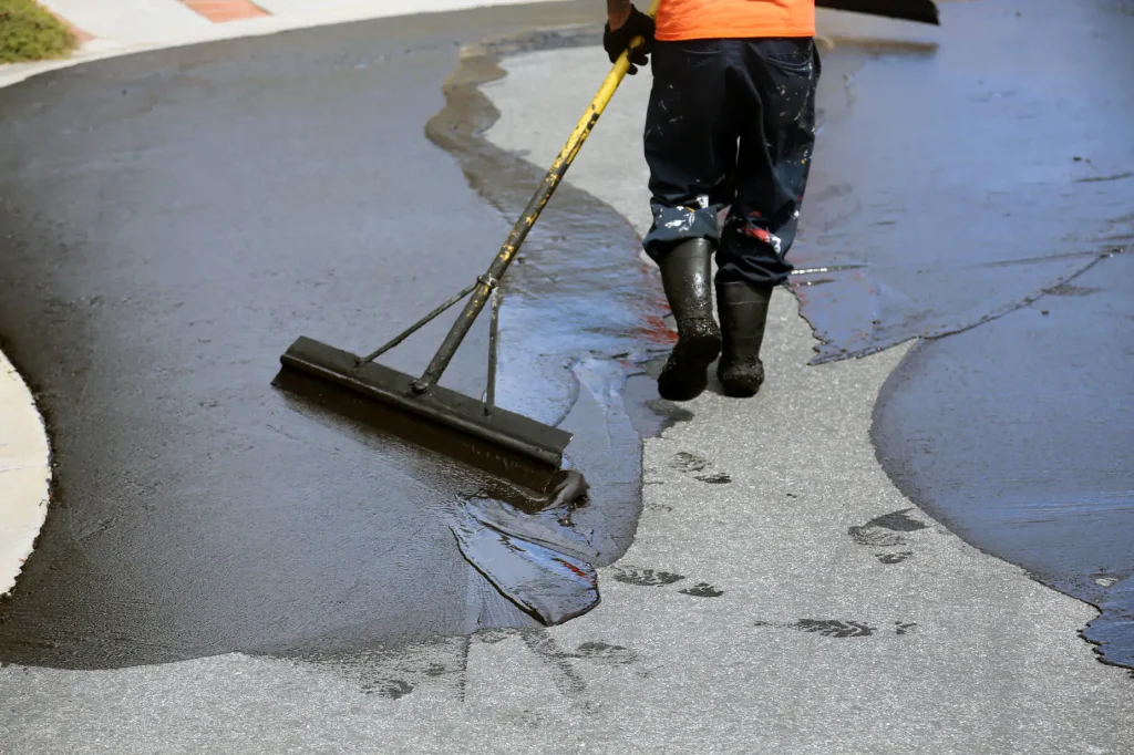 A worker in black boots and an orange shirt spreads fresh tar or sealant on a curved asphalt road using a large squeegee, leaving wet, shiny footprints behind.