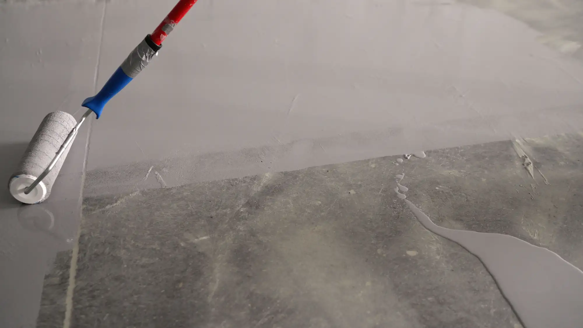 A paint roller on a handle applies a light gray coating over a concrete floor, with part of the floor already covered and part still bare.