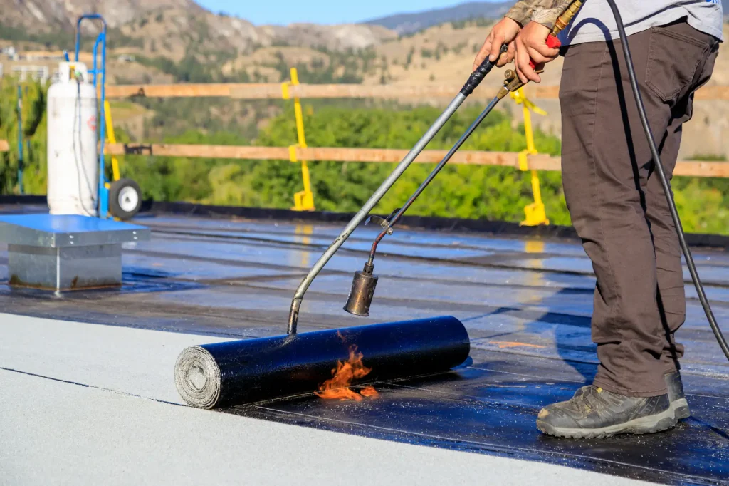 A worker uses a propane torch to apply heat to a roll of roofing material on a flat roof, fusing it in place. Construction safety barriers and trees are visible in the background.