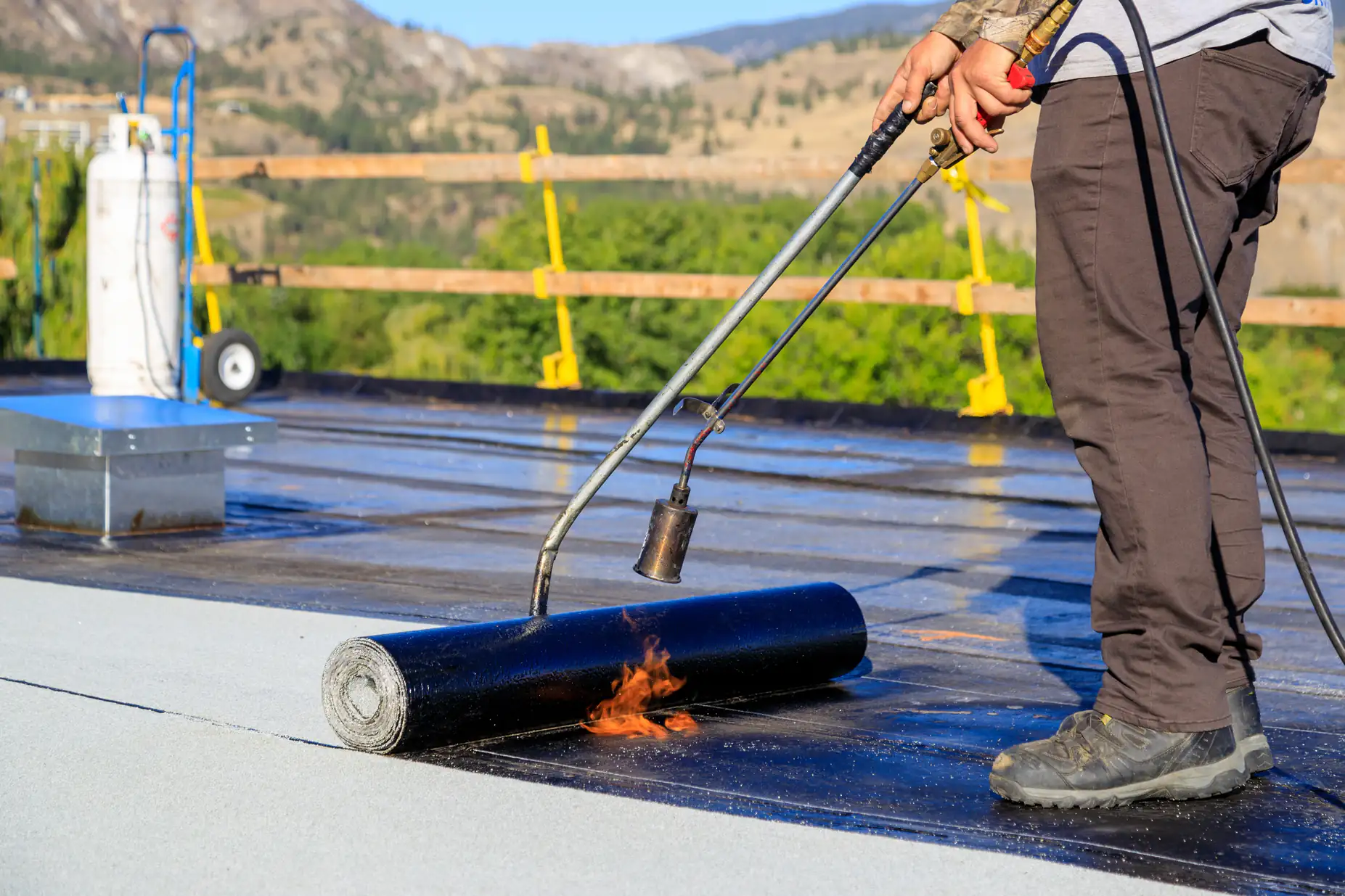 A worker uses a propane torch to apply heat to a roll of roofing material on a flat roof, fusing it in place. Construction safety barriers and trees are visible in the background.