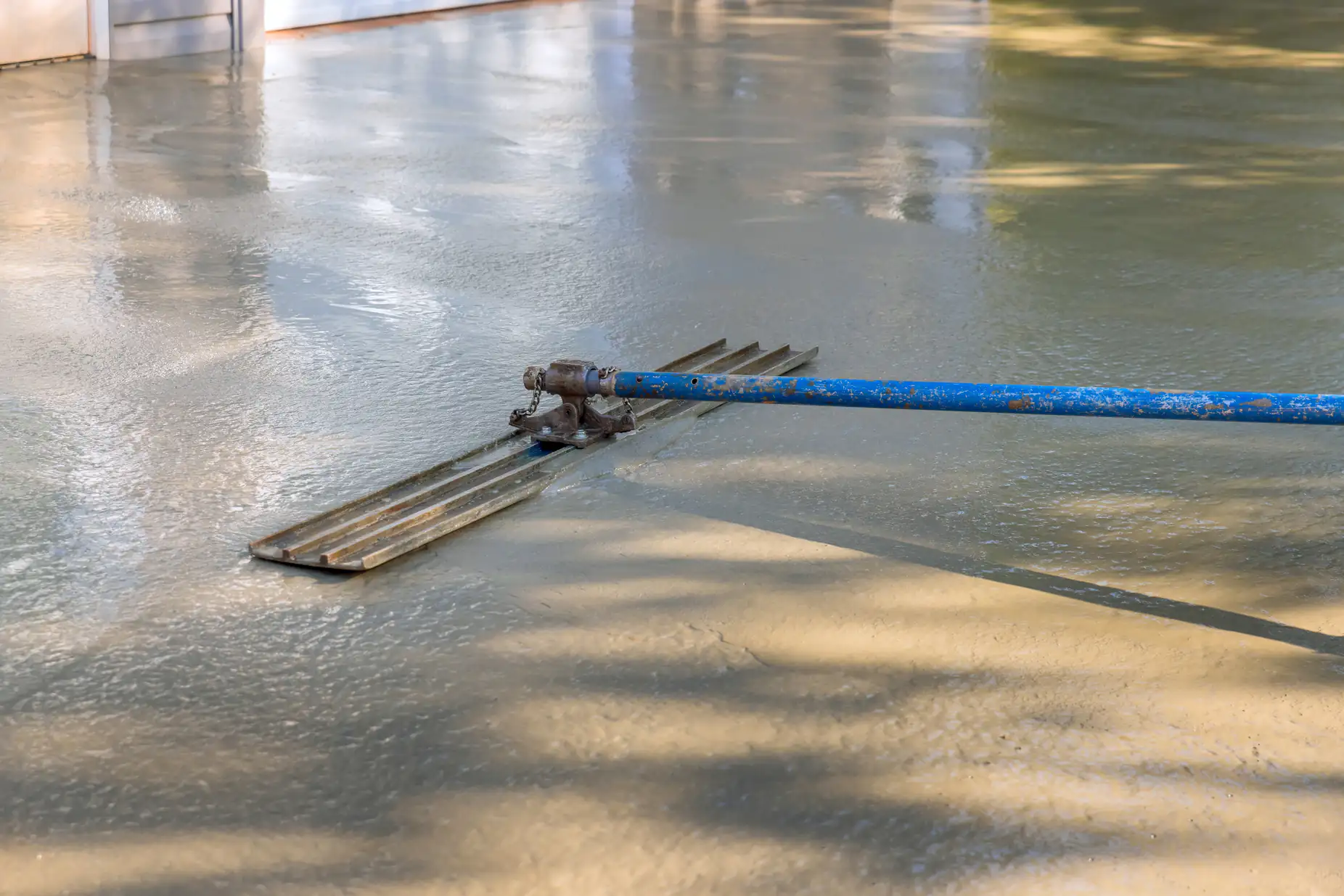 A blue-handled tool is being used to smooth and level freshly poured concrete on an outdoor surface, with reflections and shadows visible on the wet concrete.