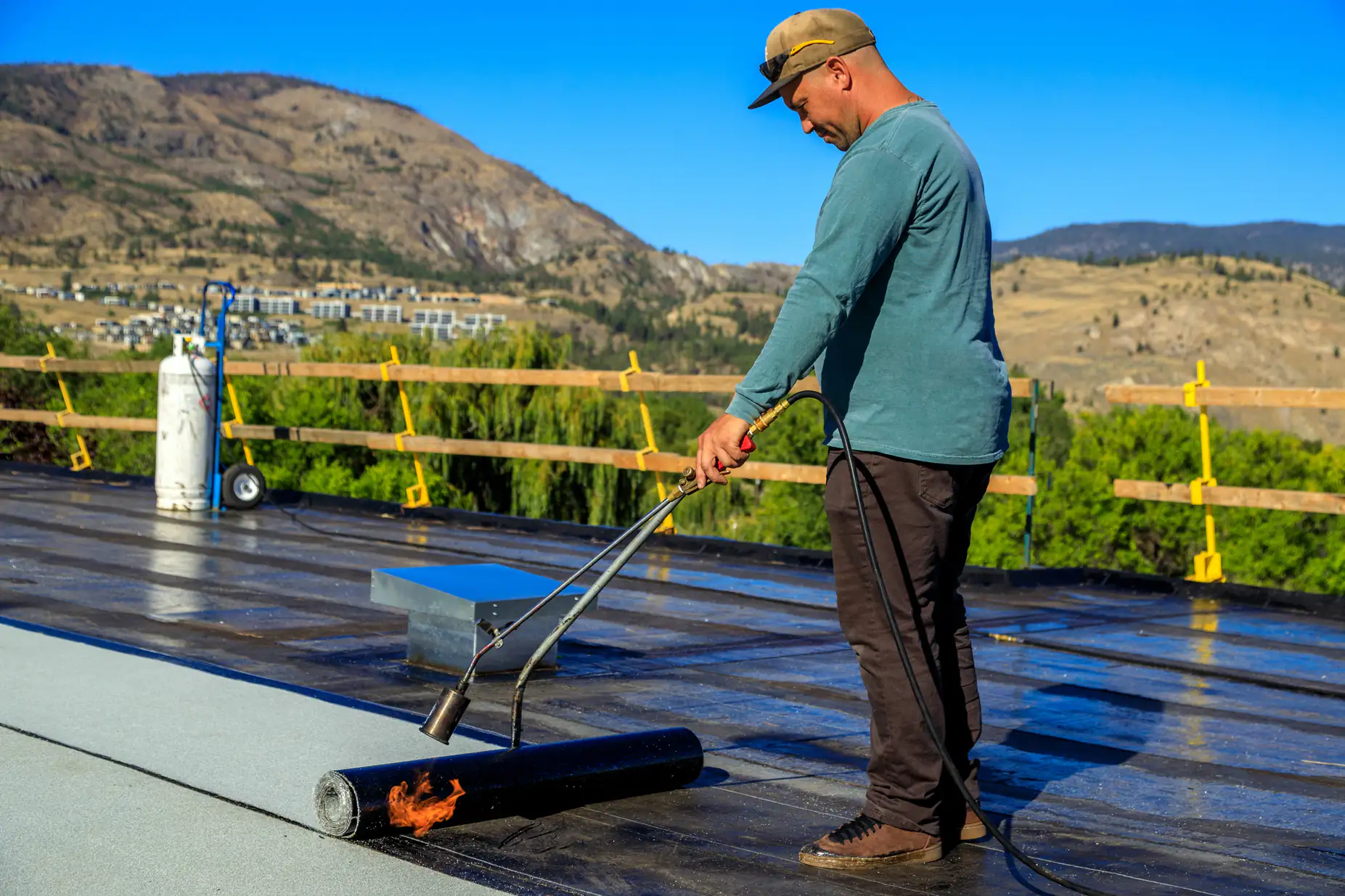 A man wearing a cap uses a torch to apply roofing material on a flat rooftop, with hills and buildings visible in the background under a clear blue sky.