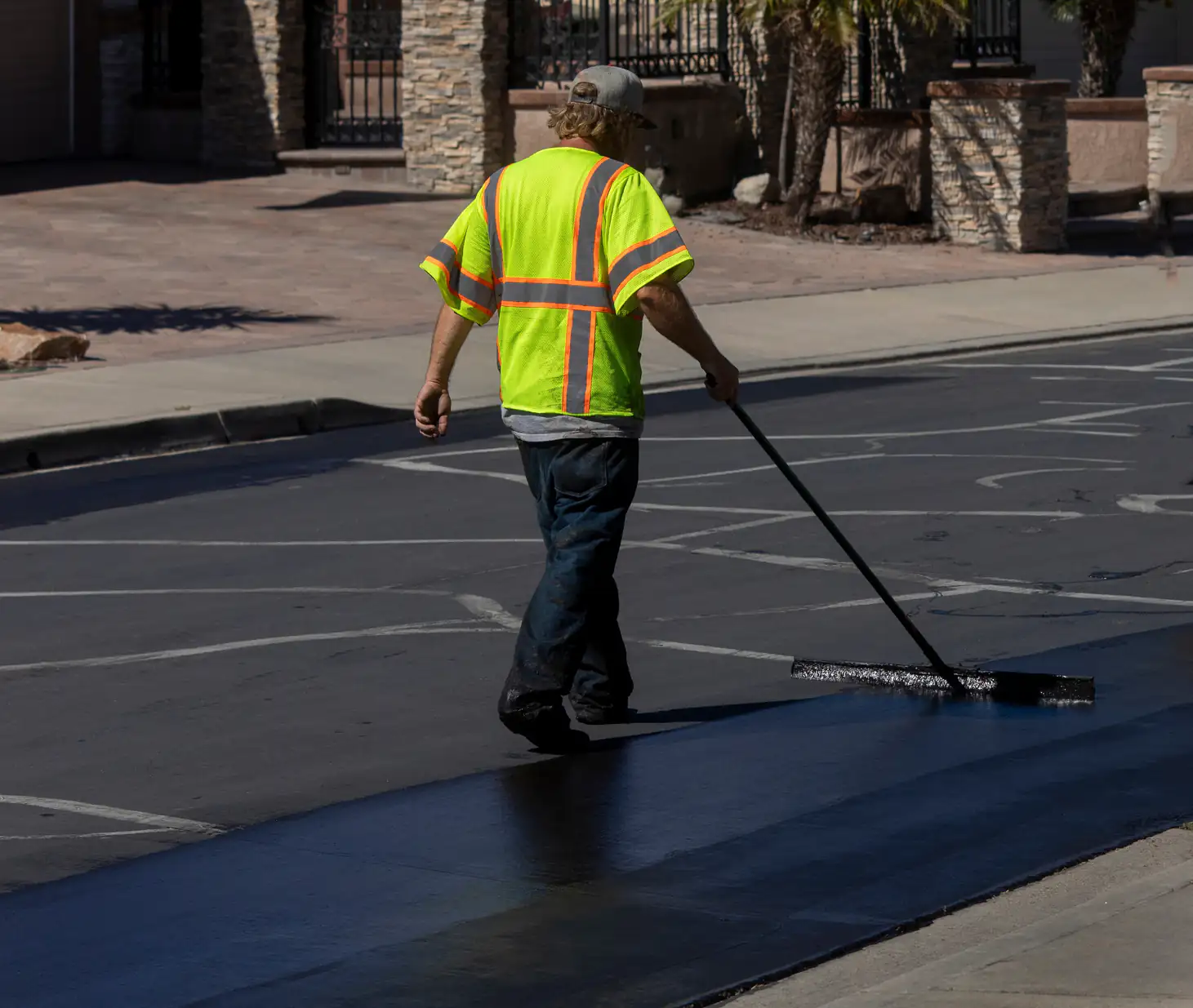 A worker in a neon yellow safety shirt and cap uses a large squeegee to spread fresh asphalt or sealant on a street in a residential area on a sunny day.
